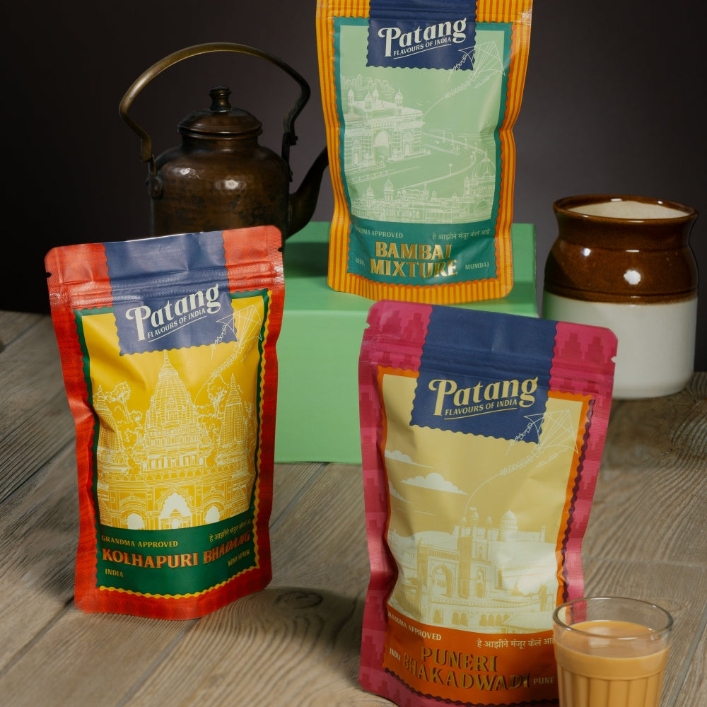 Three Patang coffee packages on a wooden surface with a glass of coffee and a teapot in the background.